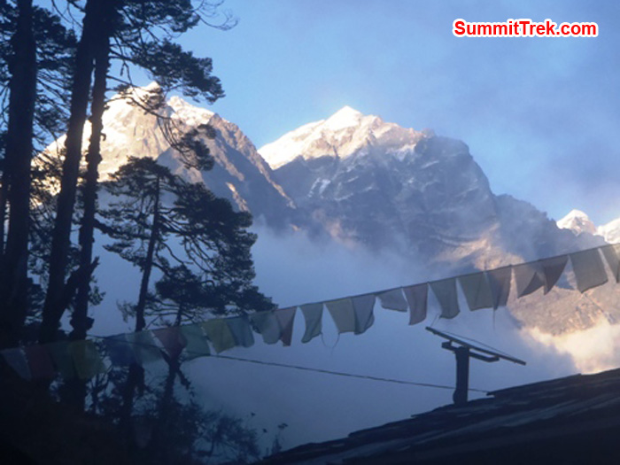 Mera Peak towers above Kote Village. In foreground prayer flags flutter and a solar panel stands upon a rooftop. Photo by Andrew Davis